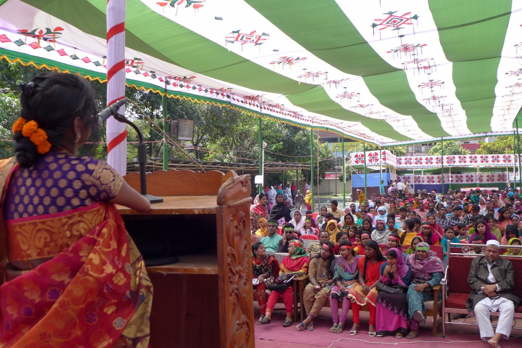 A woman wearing a sari standing a podium giving a speech to a crowd. The woman does not have hands.
